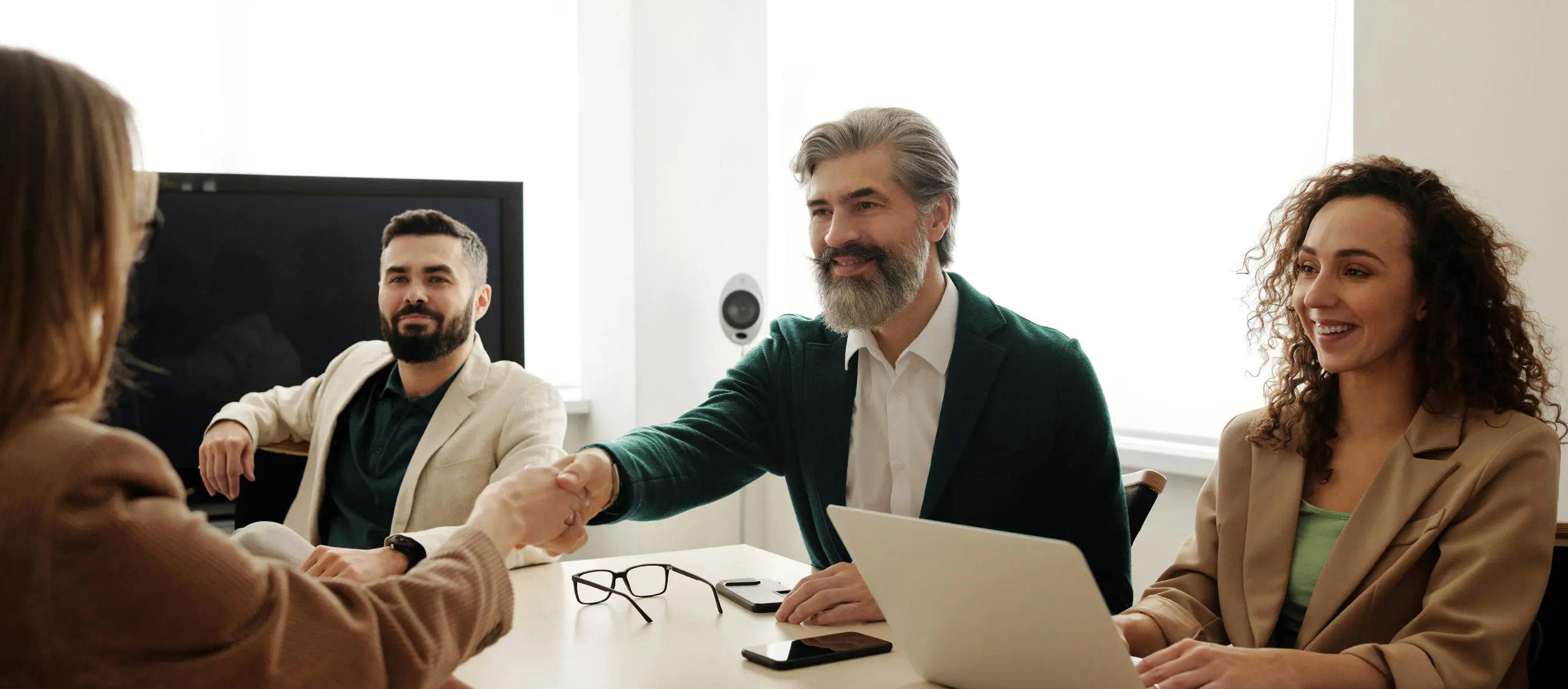 Two people shaking hands across a desk during a meeting with colleagues, symbolizing a professional agreement.